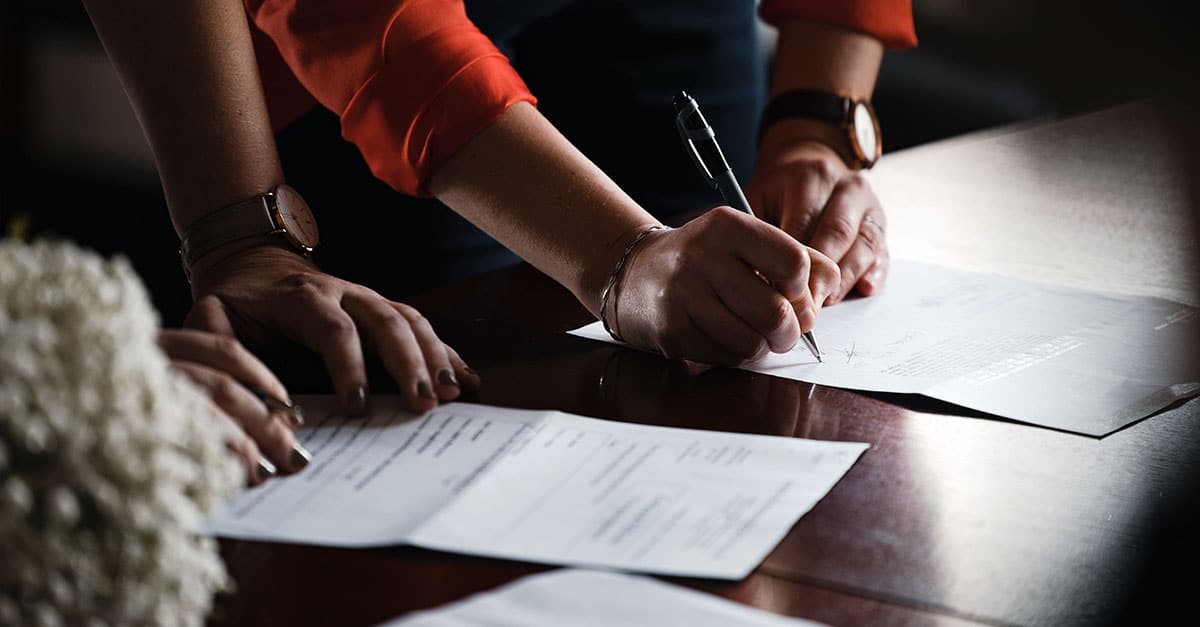 Image of two pairs of hands, one is signing an agreement and one touching the agreement. Image is used to illustrate of signing a shareholders' agreement.